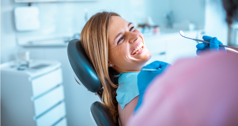 Woman sitting in dentist chair in clinic room laughing while dentist is about to treat her teeth.