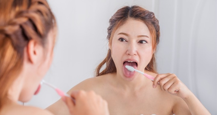 Woman cleaning her tongue with her toothbrush in front of a mirror in the bathroom.