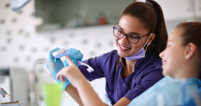 Dental hygienist educating a young female patient on how to use a toothbrush to clean her braces.