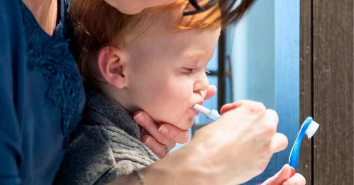 A mother brushing her toddler’s teeth while he holds on to another toothbrush