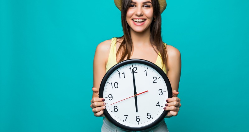 A smiling young woman holds a clock while asking how long does invisalign take to straighten my teeth.
