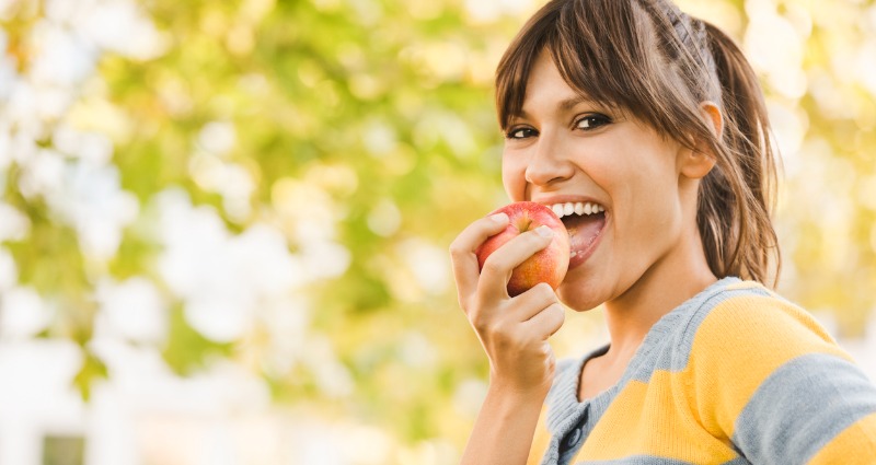 A young woman smiling at the camera and biting into an apple because she has strong teeth.